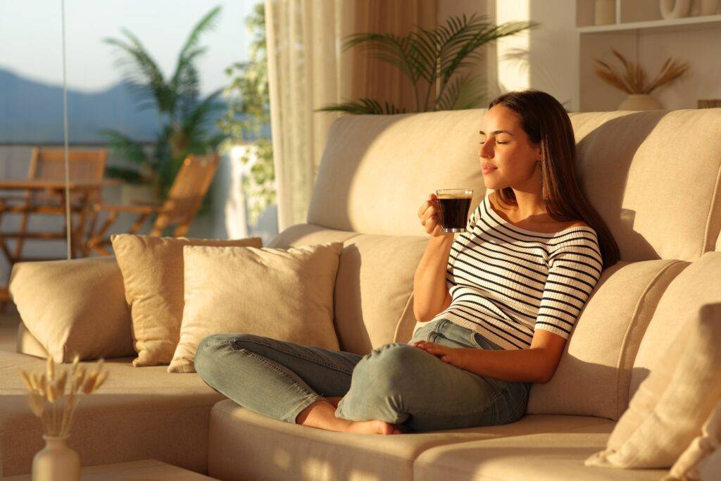 Frau genießt Kaffee auf Sofa im warmen Sonnenlicht | Bettbank
